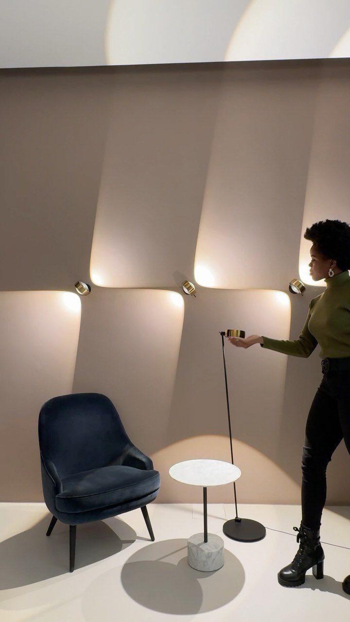A woman reaches for a minimalist floor lamp near a blue velvet chair and marble table.