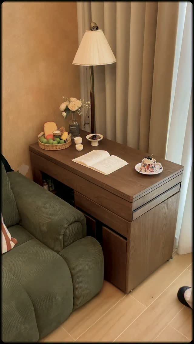 A wooden console table holding a lamp, fruit, and an open book next to a green sofa.