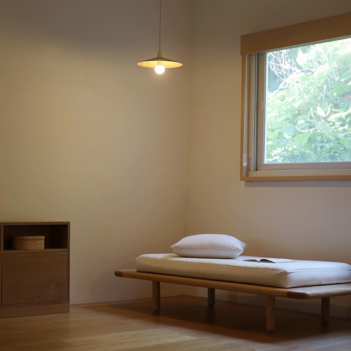 A minimalist wooden daybed with a white mattress and pillow in a bright room.