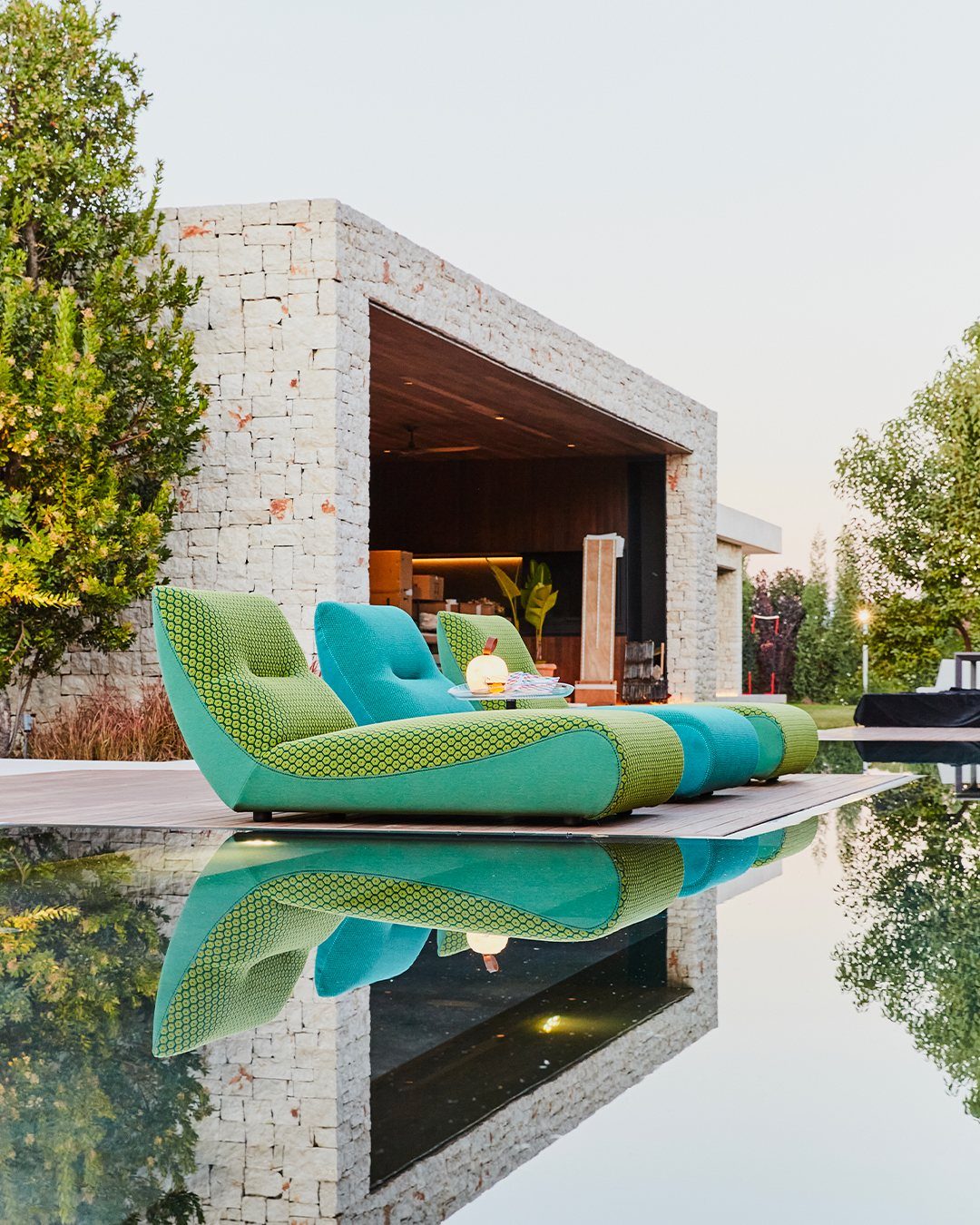 Two curved green lounge chairs sit on a wooden deck next to a calm reflecting pool.