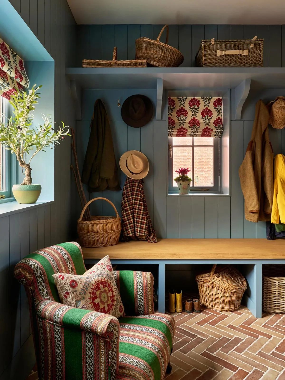 Blue mudroom with wooden bench, wicker baskets, hanging coats, and a patterned green armchair.
