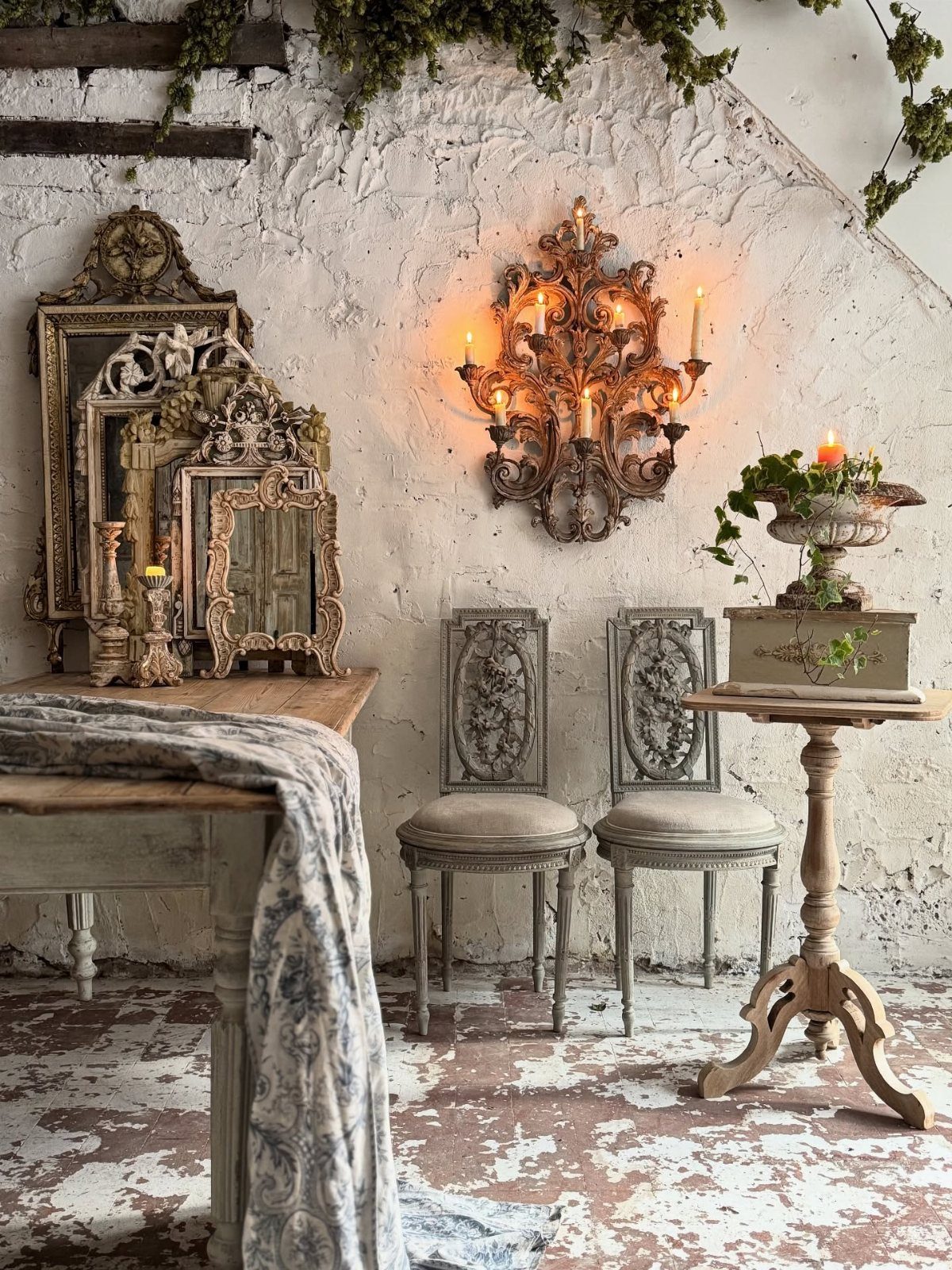 Ornate vintage mirrors, carved chairs, and a wrought iron candelabra against a textured white wall.