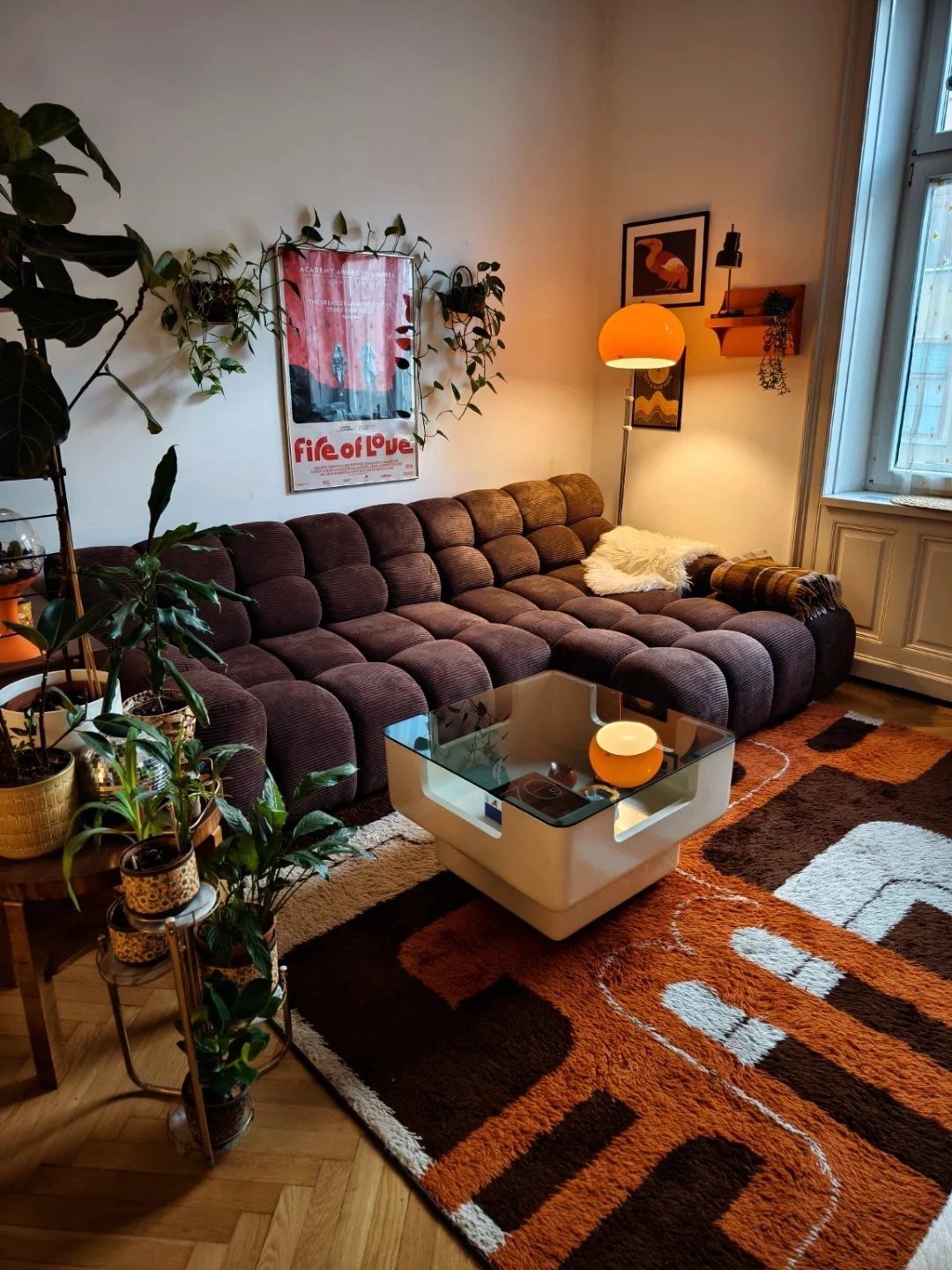 A brown tufted sofa sits near a glass coffee table and patterned rug surrounded by plants.
