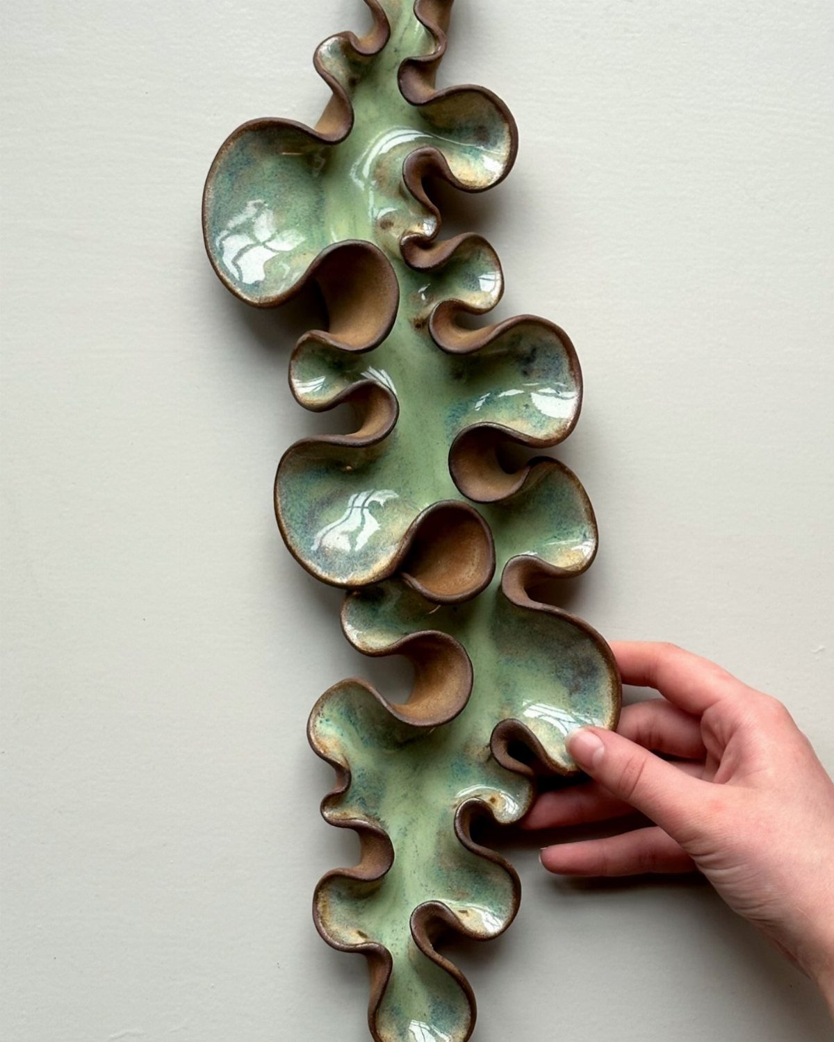 A hand touching a large, wavy teal and brown glazed ceramic wall sculpture.