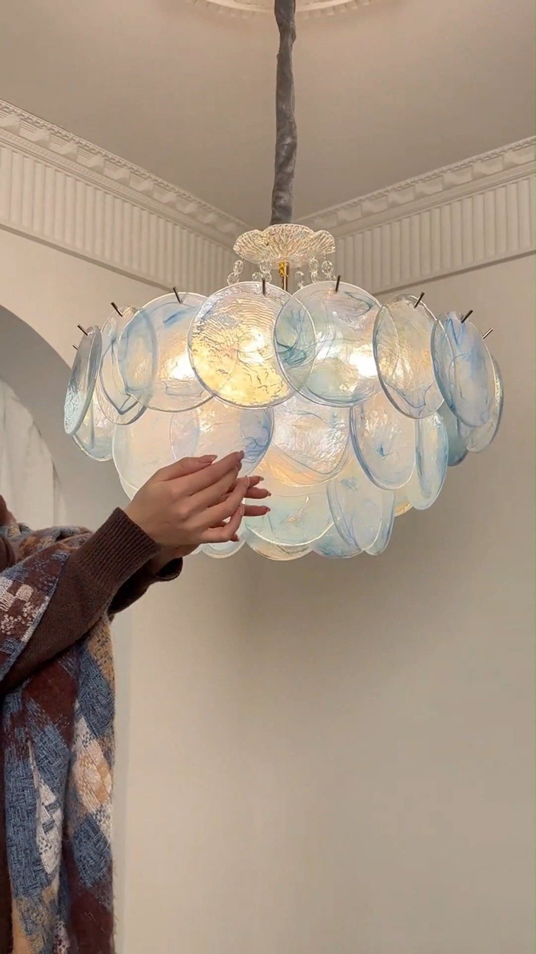 A hand reaching toward a large, iridescent circular glass disc chandelier hanging from a white ceiling.