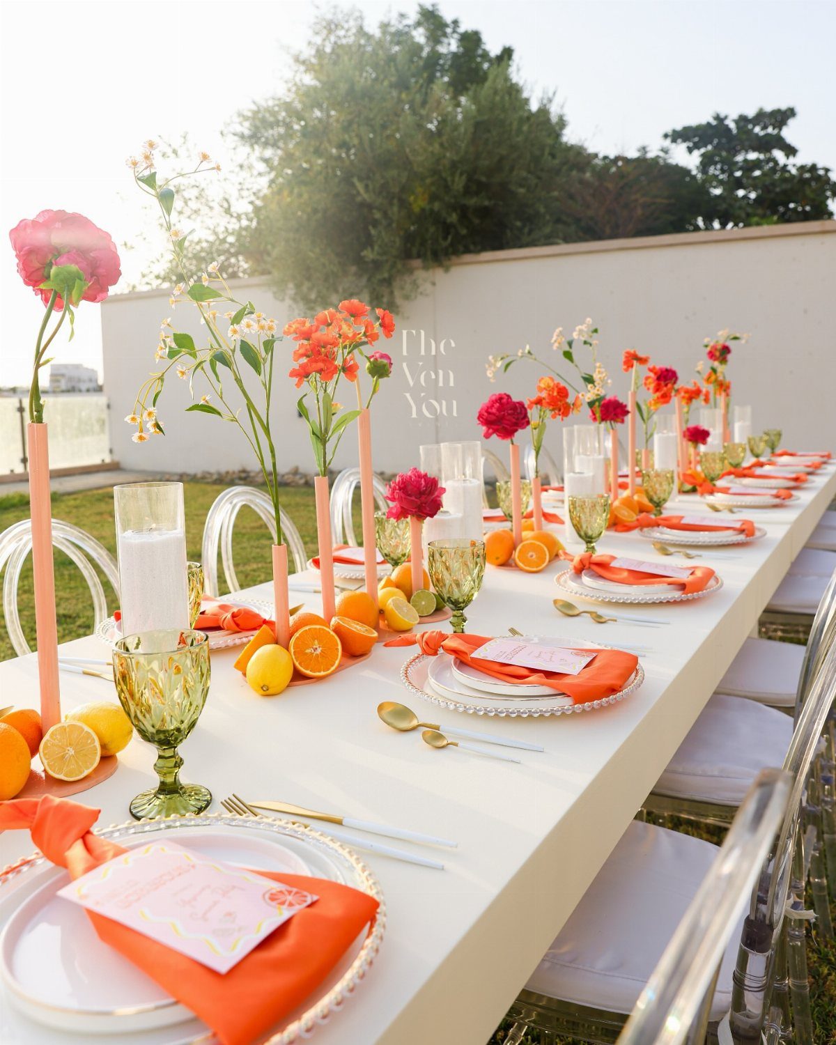 A long white table decorated with orange linens, citrus fruits, and tall pink floral vases.