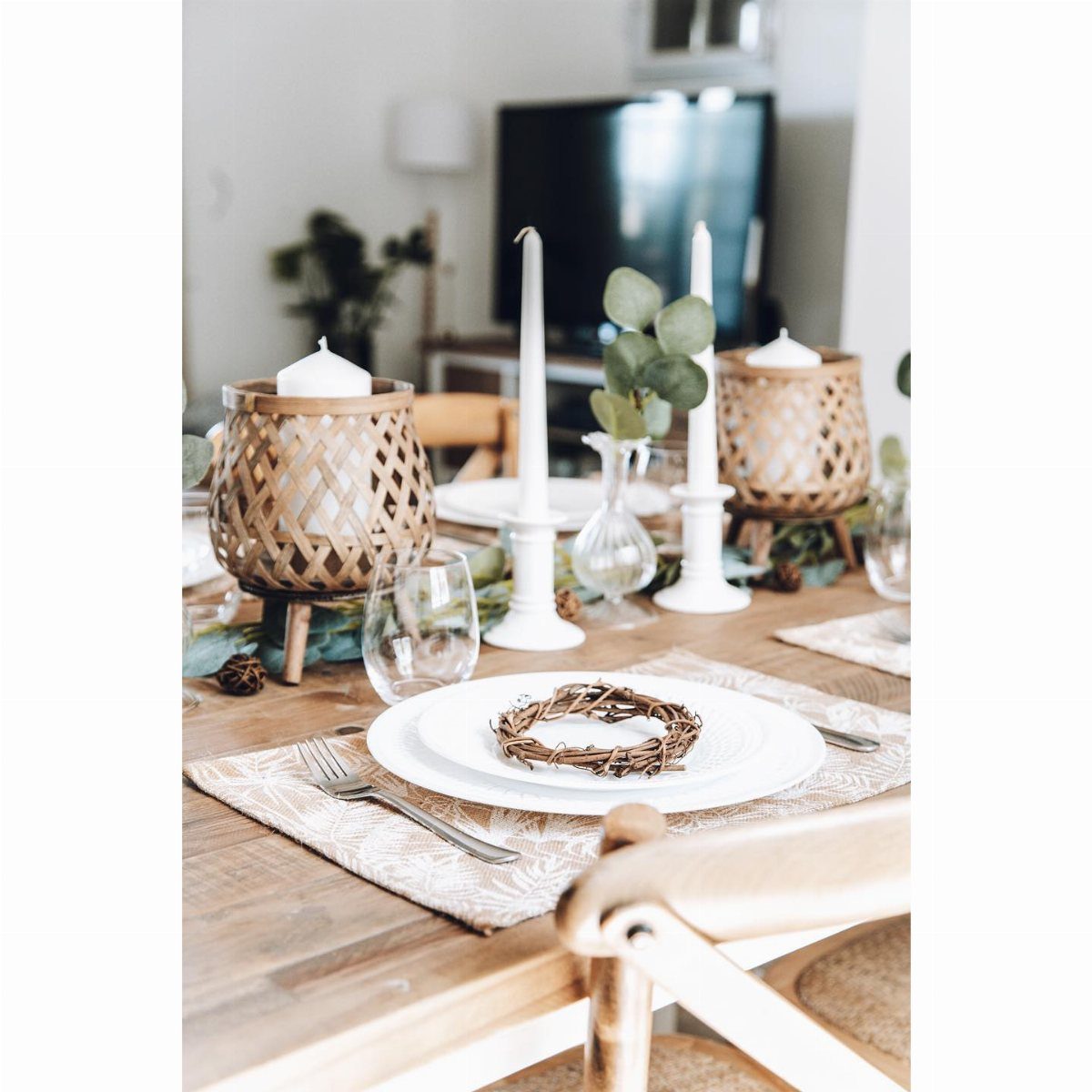 A wooden dining table set with white plates, woven candle holders, and a twig wreath centerpiece.
