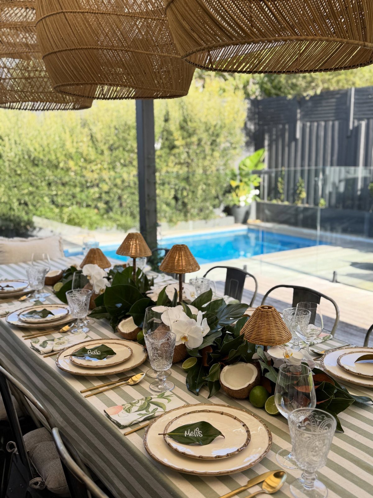 A long outdoor dining table set with striped linens, greenery, and coconut decor by a pool.