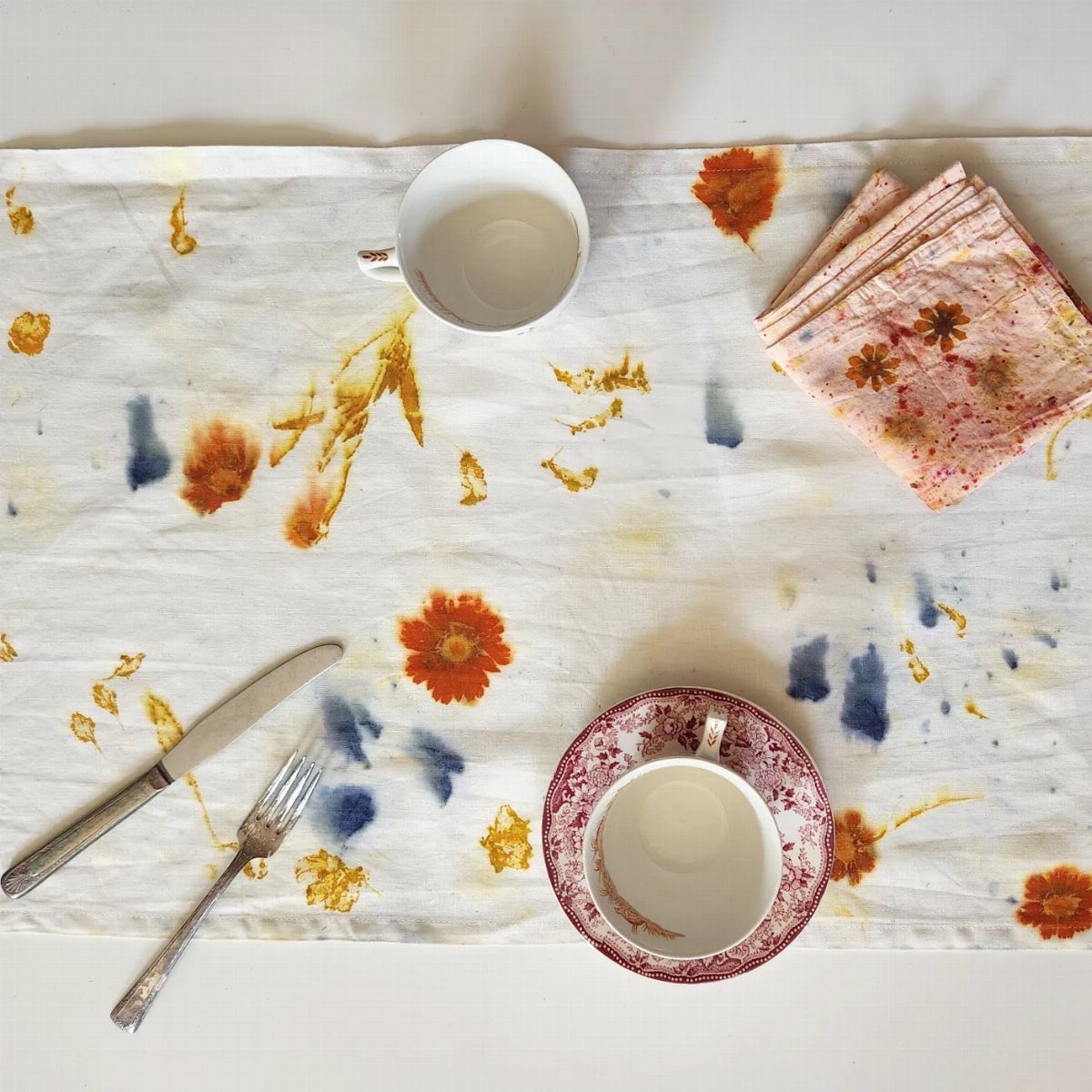 White table runner with colorful organic floral patterns, teacups, silverware, and folded napkins on a white surface.