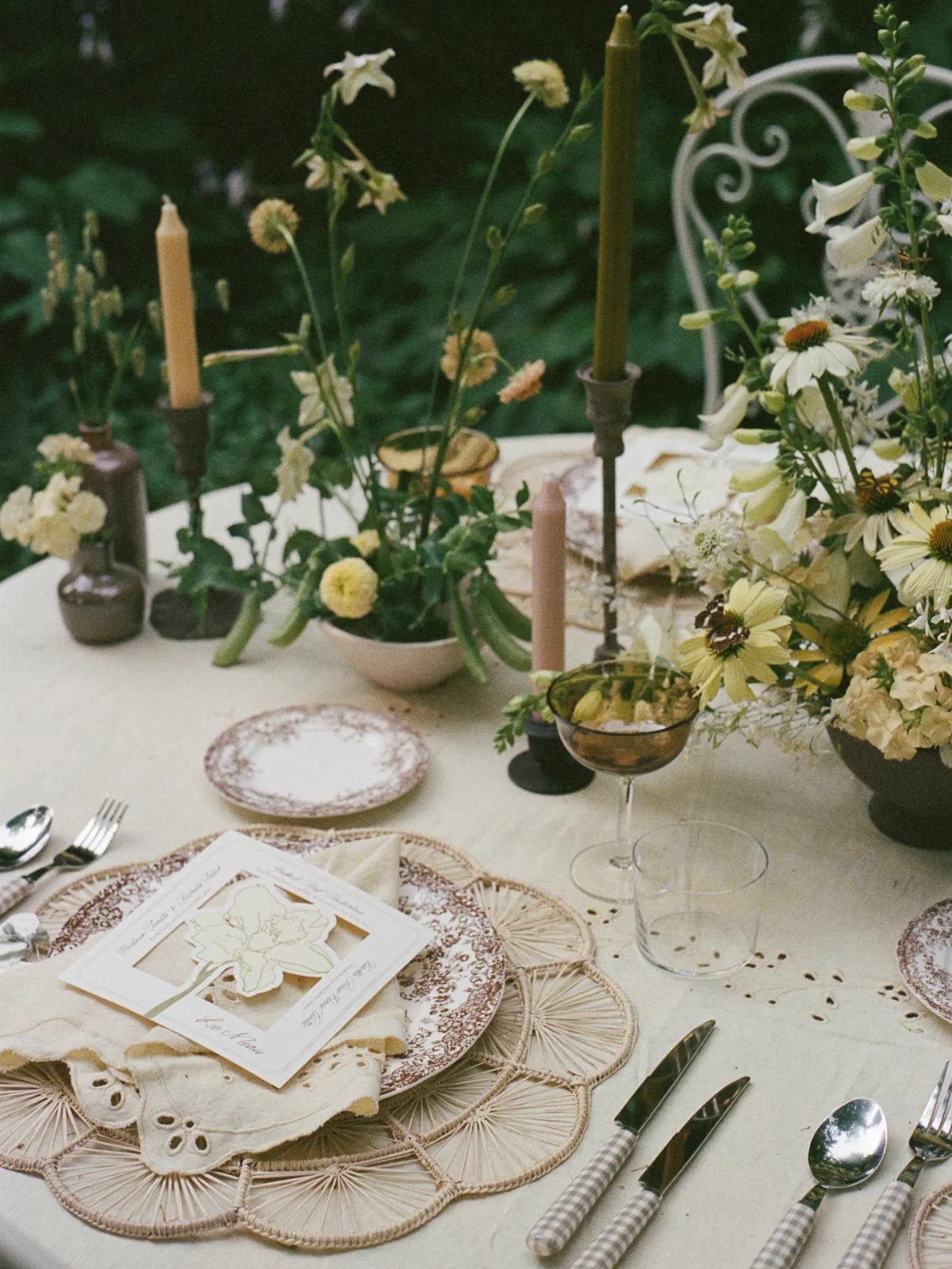 A summer table setting with eyelet linens, patterned plates, woven placemats, and yellow flowers.