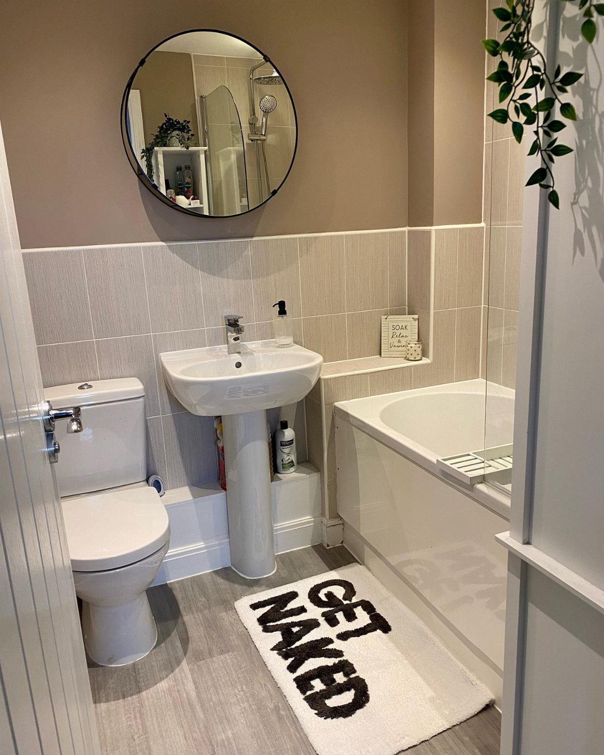 A small bathroom with white fixtures, taupe tiled walls, and a round mirror.