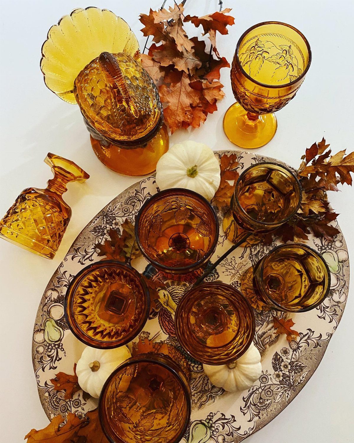 Amber vintage glassware and small white pumpkins arranged with autumn leaves on a patterned platter.