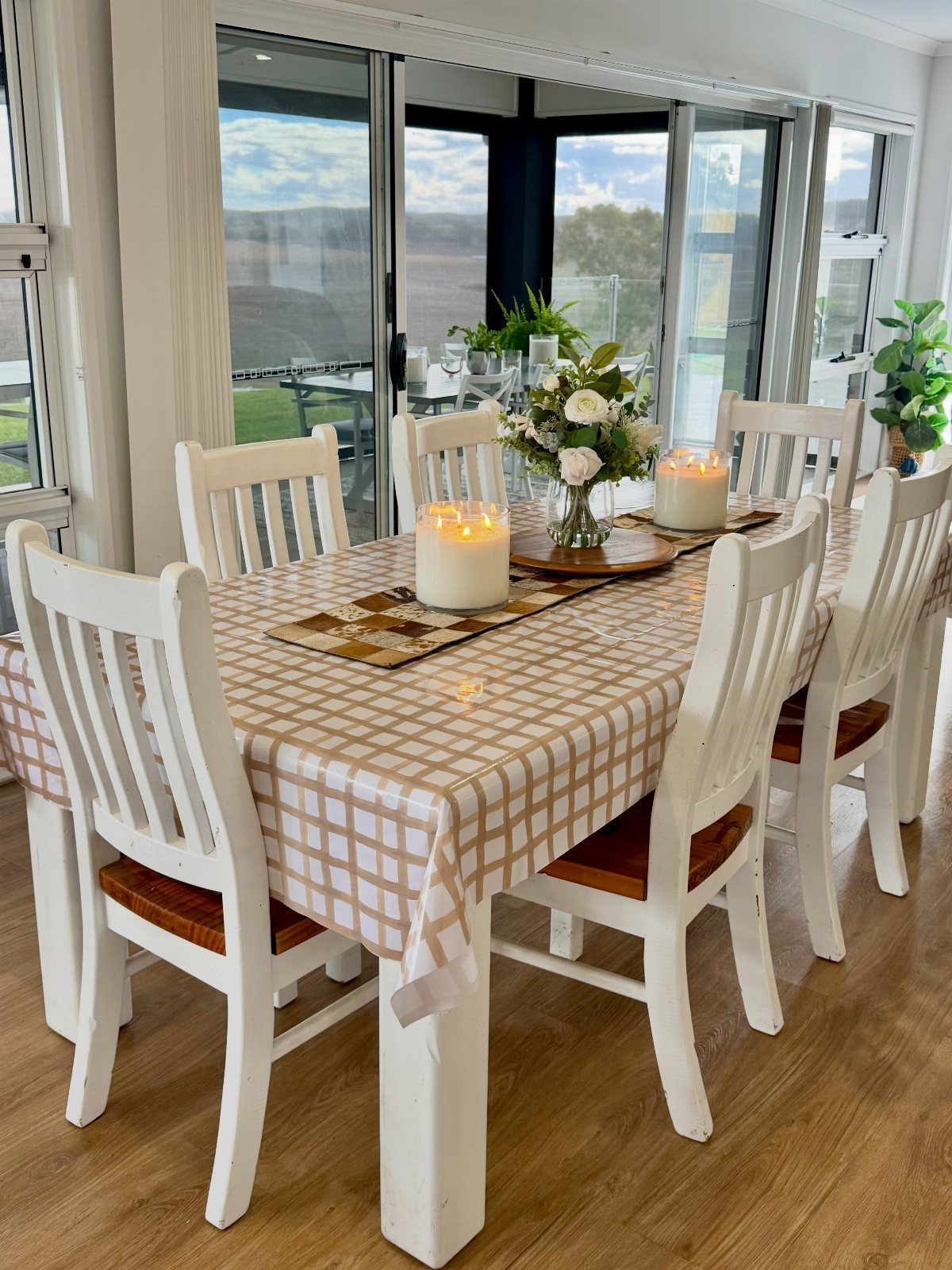 A dining table set with a checkered tablecloth, candles, and flowers near large glass doors.