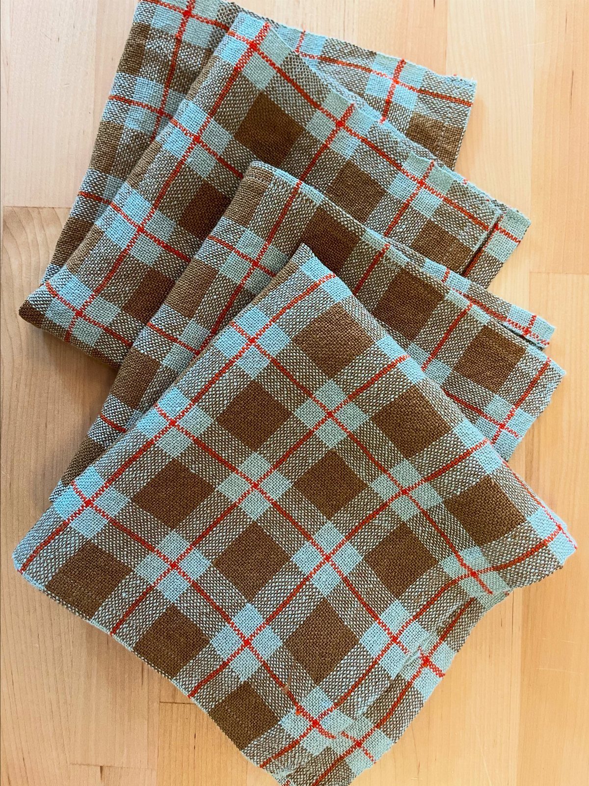 Plaid fabric napkins with brown, blue, and red patterns arranged on a light wooden tabletop.