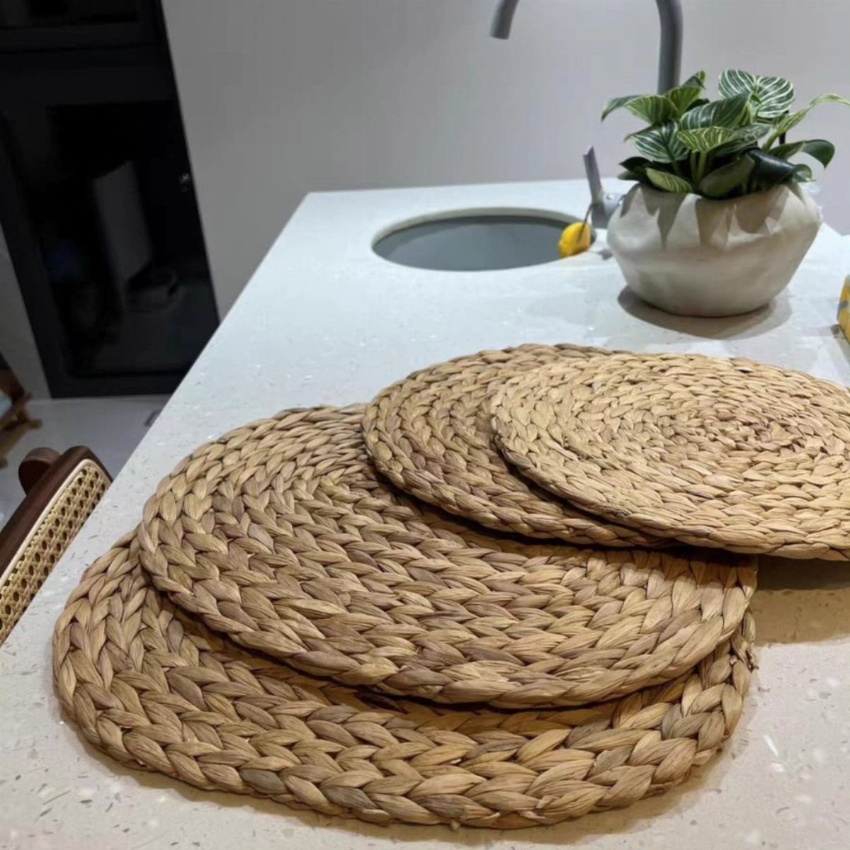 Round woven straw placemats stacked on a white kitchen countertop near a sink.