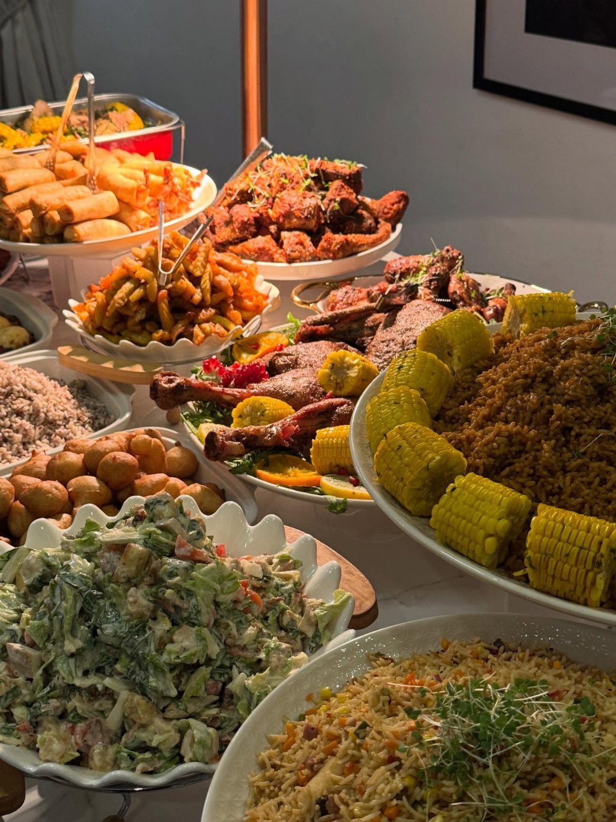 A large buffet spread with corn, fried chicken, rice, and various salads in white serving dishes.