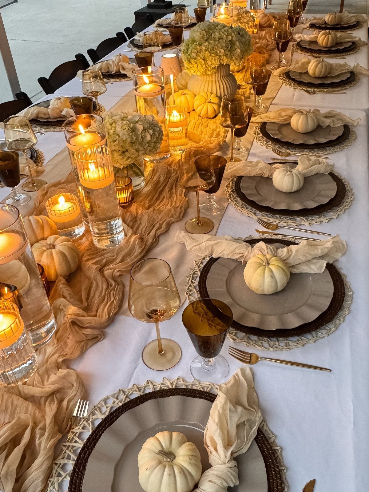 A long dining table featuring layered plates, white pumpkins, lit candles, and a tan fabric runner.