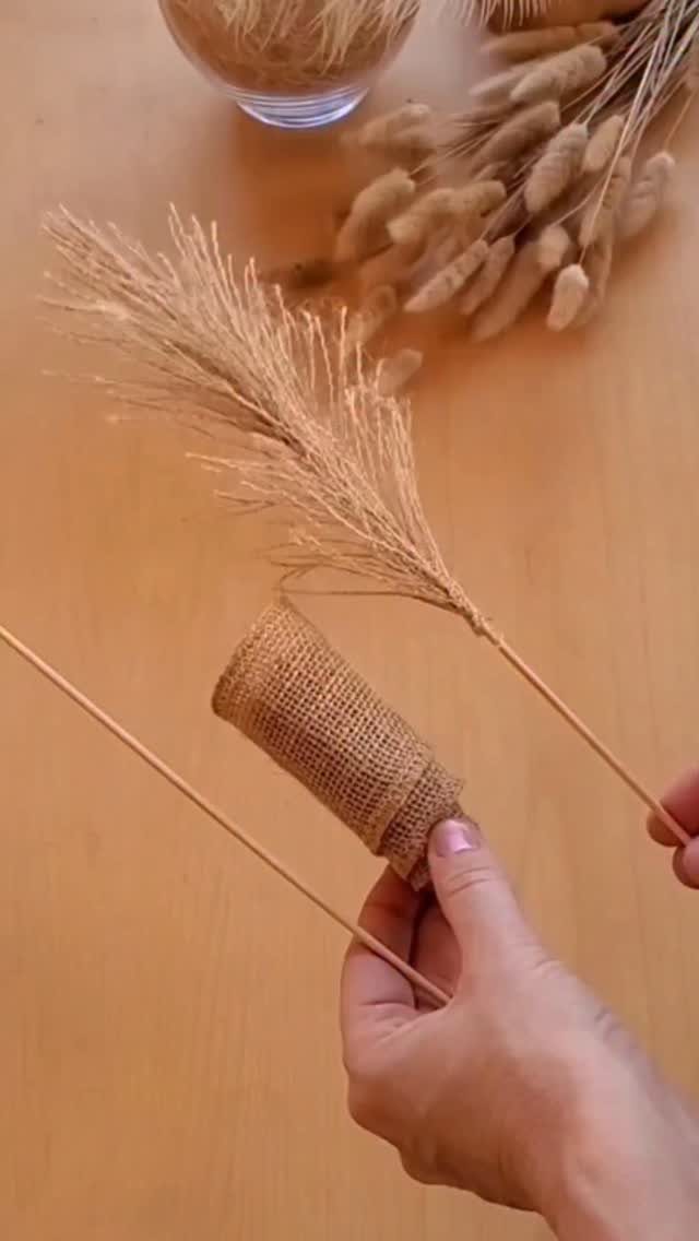 Hands wrapping a wooden stick with burlap next to dried pampas grass stems.