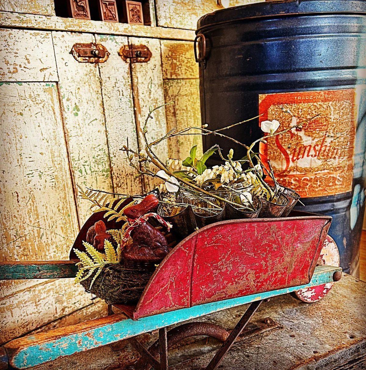 Small red rustic wheelbarrow filled with dried branches and white flowers on a wooden surface.