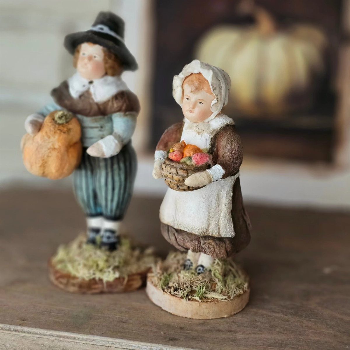 Two small pilgrim figurines holding a pumpkin and a fruit basket on a wooden surface.