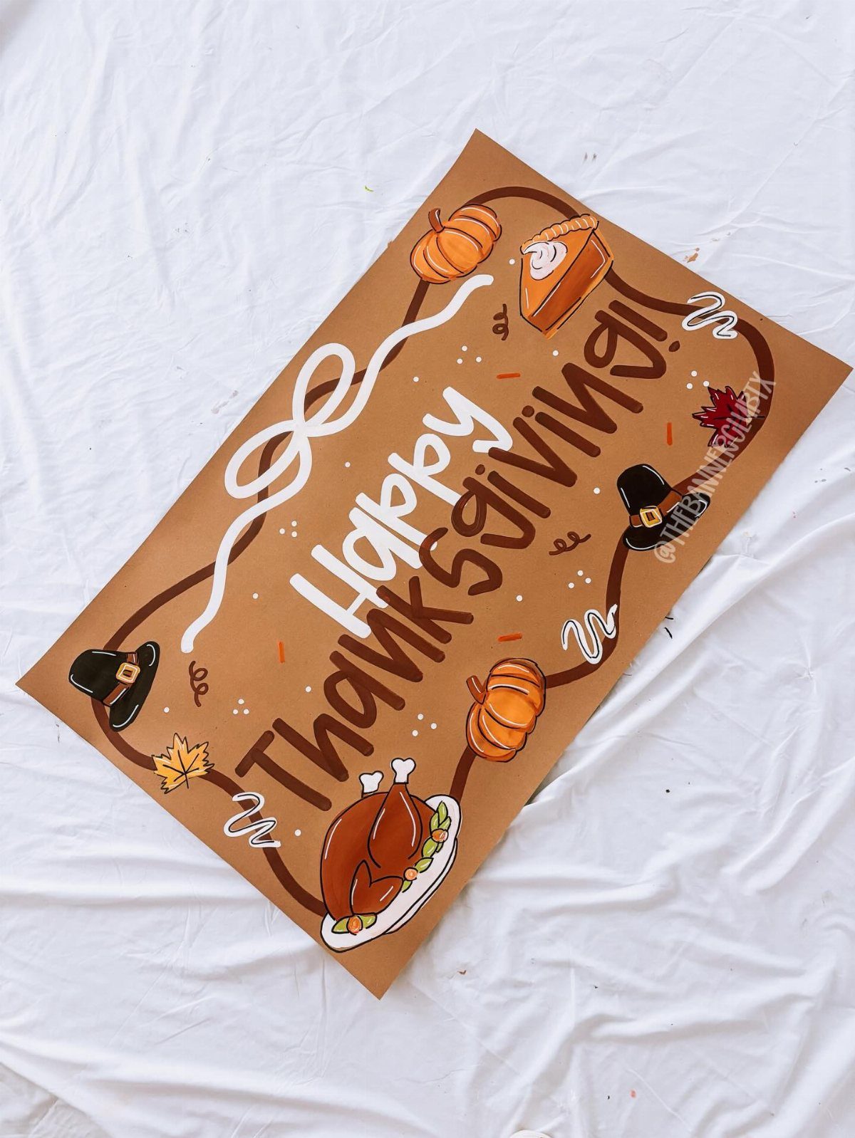 Hand-painted brown Thanksgiving sign with turkey, pumpkin, and pilgrim hat illustrations on white fabric.