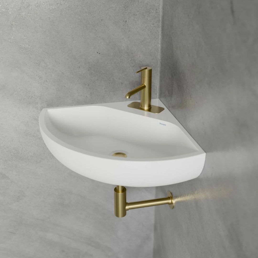 White triangular corner sink with a gold faucet mounted against grey tiled walls.