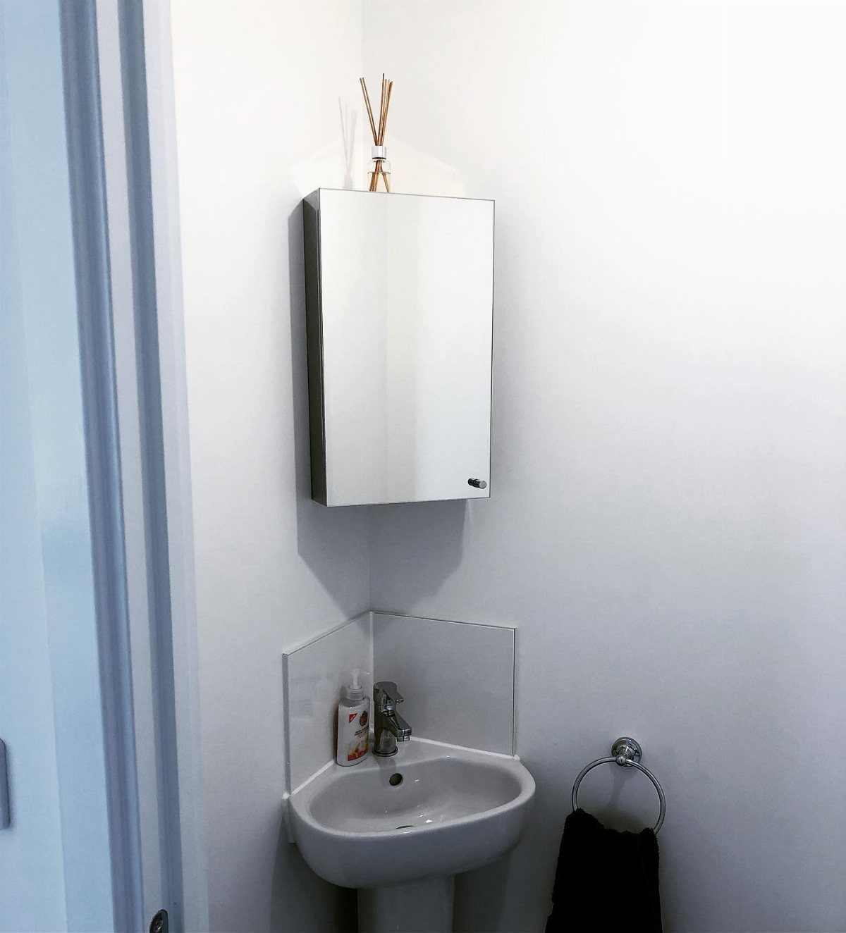 White corner sink with chrome faucet and a mirrored medicine cabinet on a white wall.