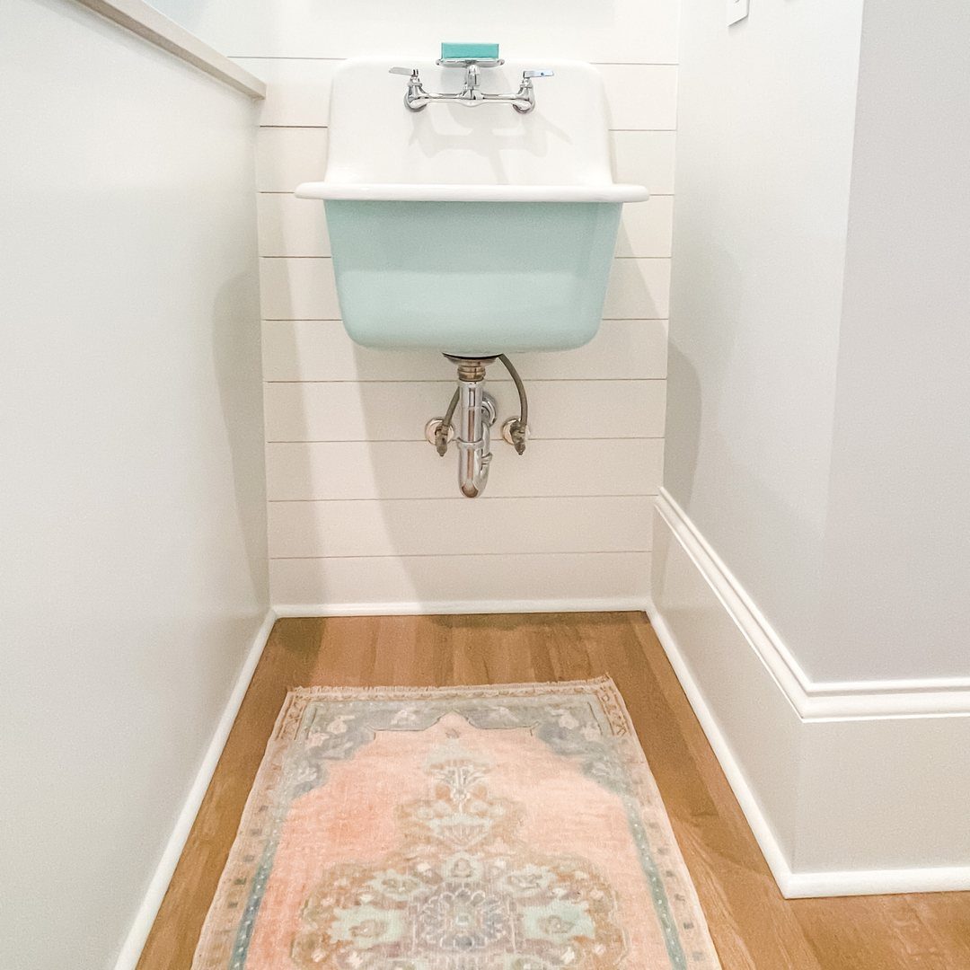 Light blue wall-mounted sink with silver faucets against white shiplap walls and a patterned rug.