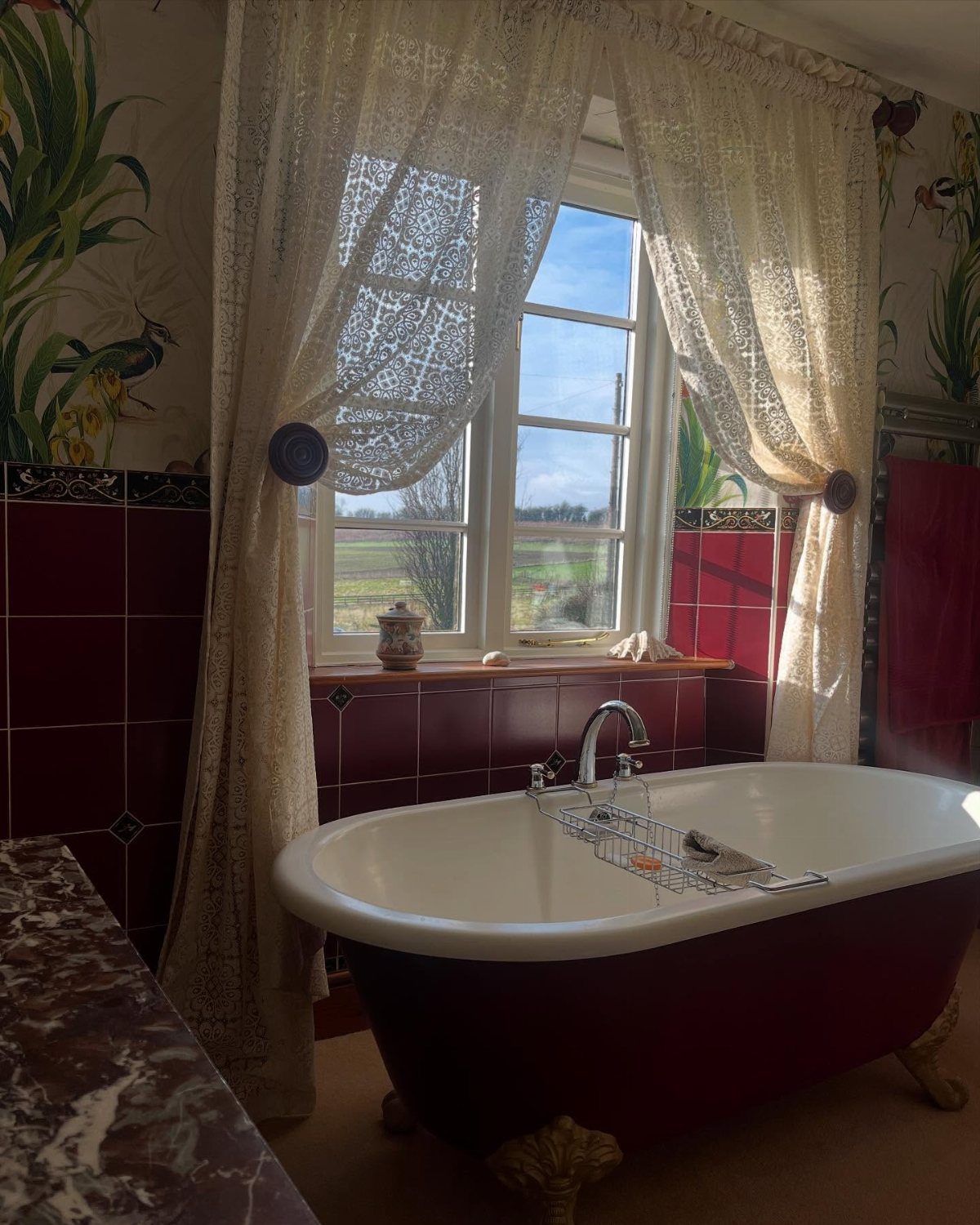 Cream lace curtains hanging over a window next to a red and white clawfoot bathtub.