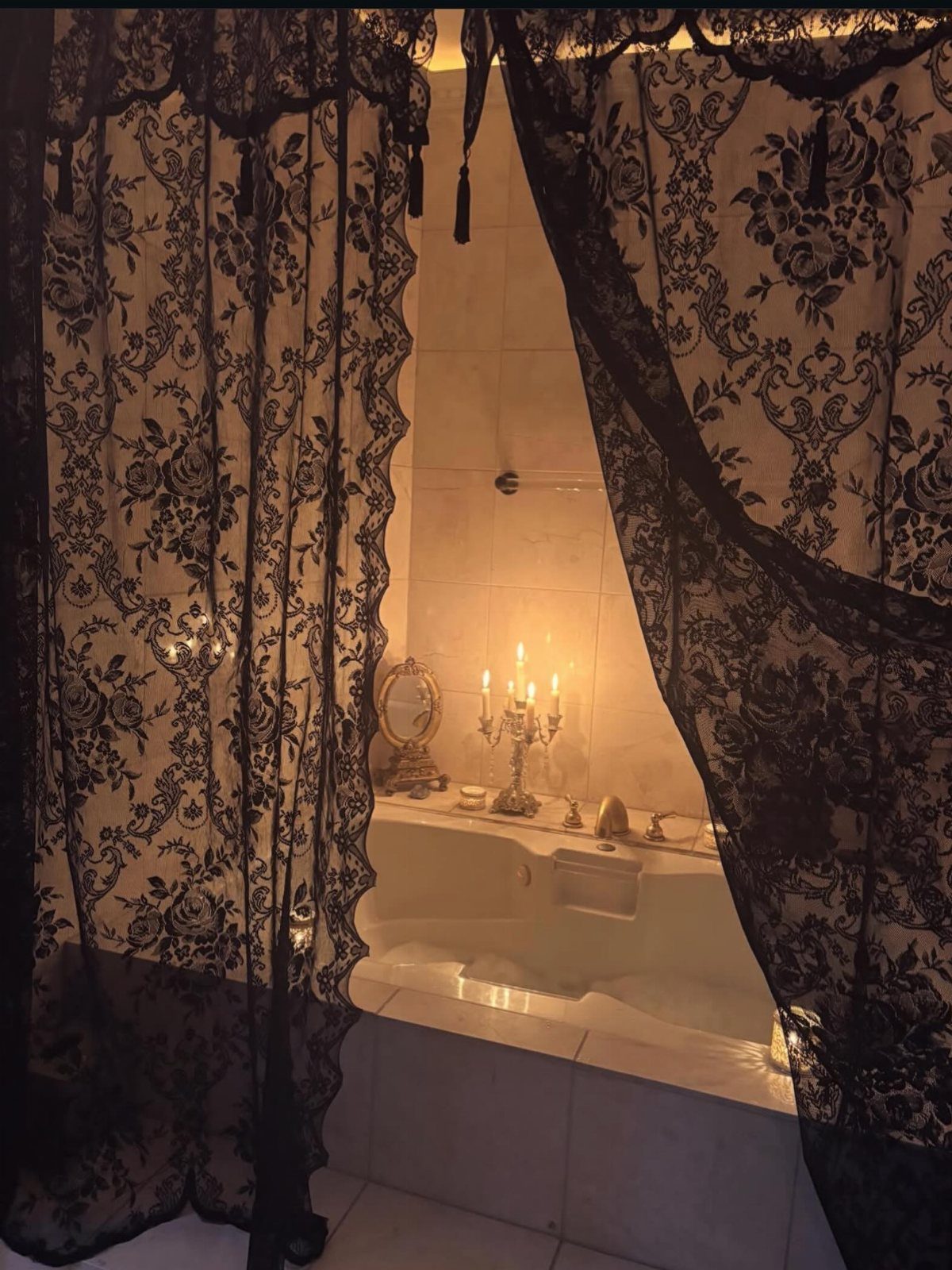 Lit candles in a silver candelabra beside a bubbly bathtub framed by black lace curtains.
