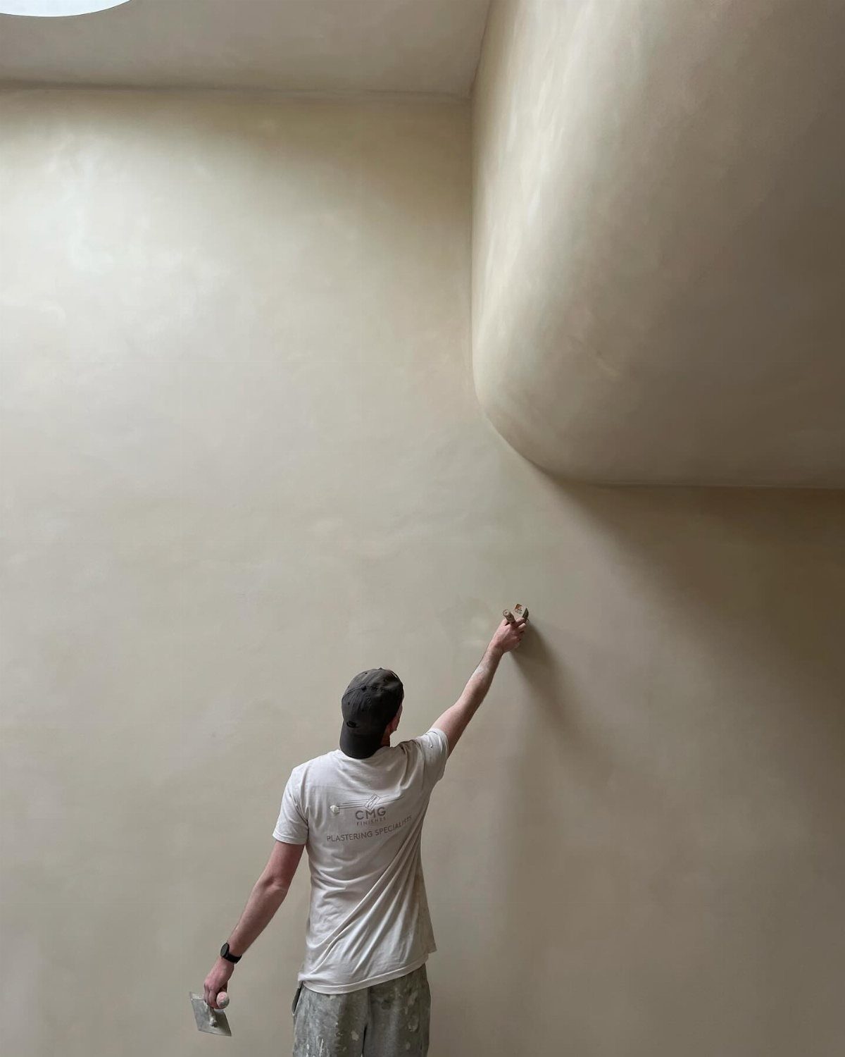 A worker applying smooth beige microcement finish to a large curved wall with a trowel.
