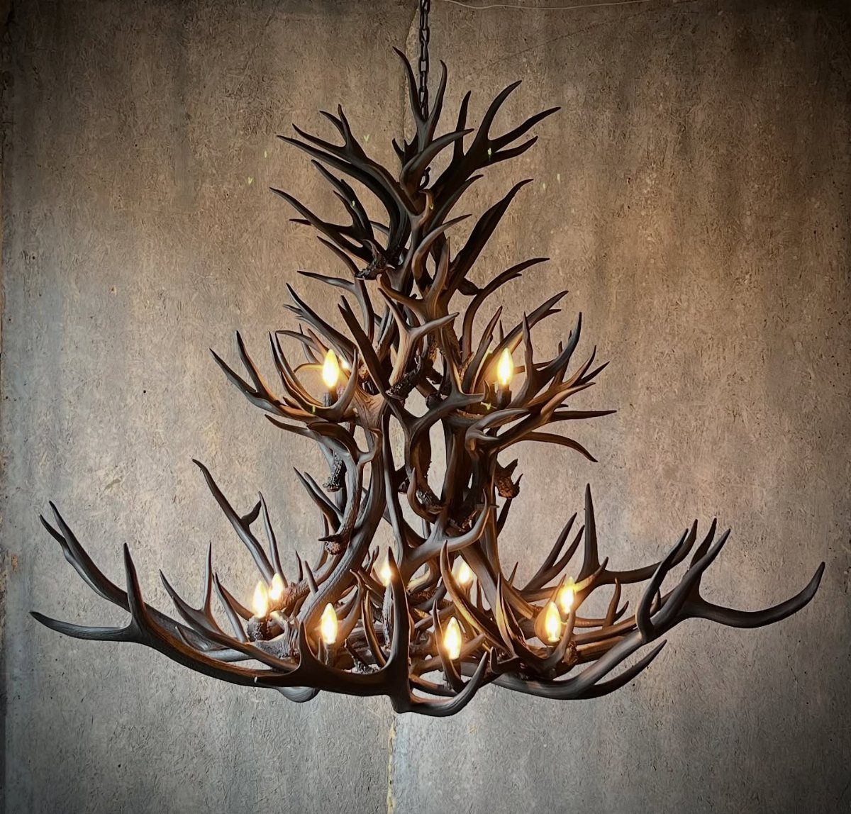 A rustic antler chandelier with warm glowing lights hanging against a grey textured wall.