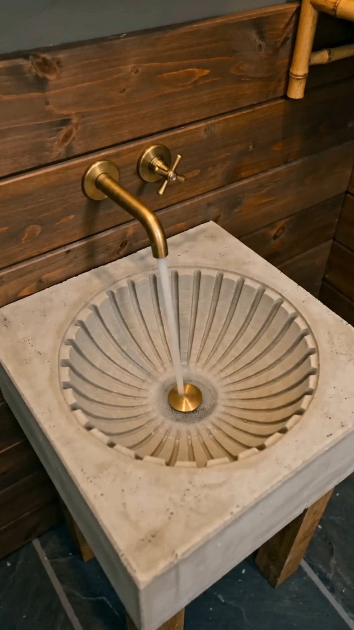 A fluted stone basin with running water from a brass wall-mounted faucet against wood paneling.