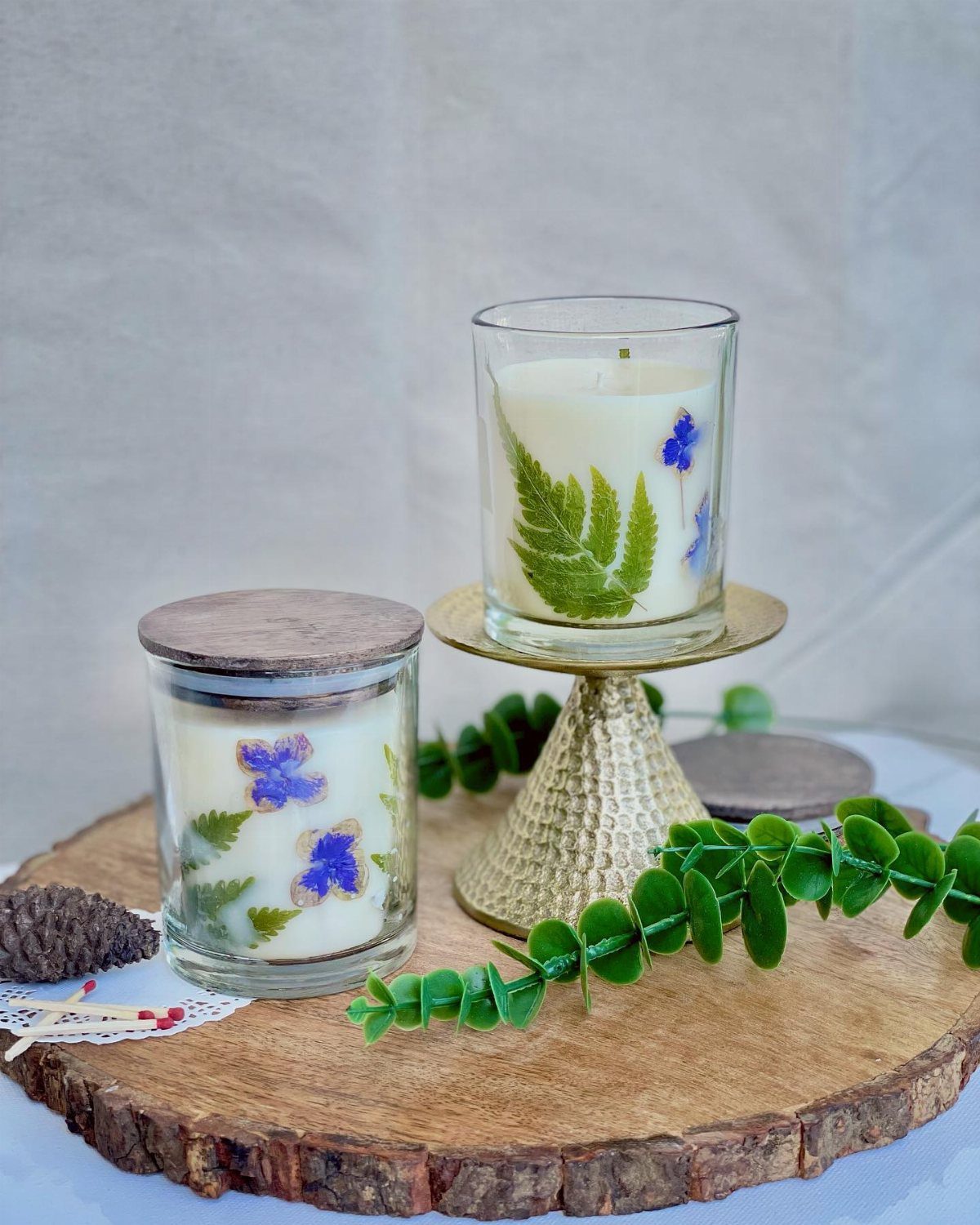 Two decorative candles with pressed botanical leaves and flowers on a wooden slab.