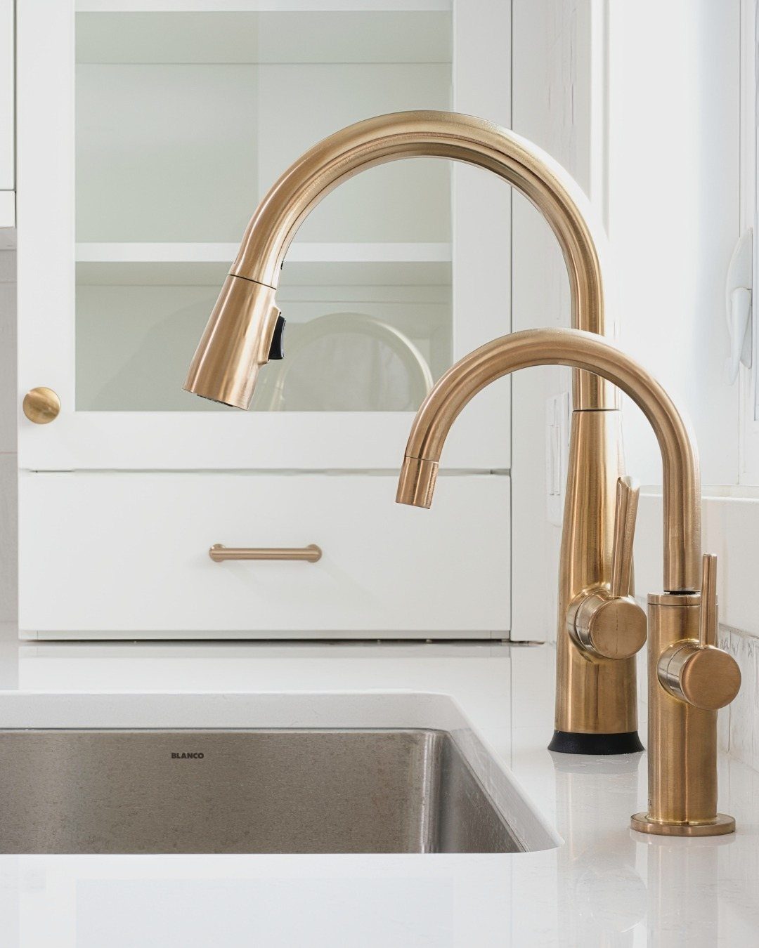 Brushed gold kitchen faucets installed on a white countertop next to a stainless steel sink.