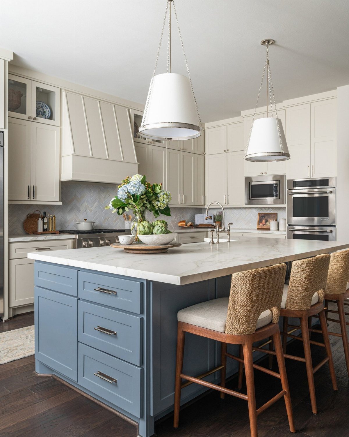 Blue kitchen island with marble countertop, woven bar stools, and white pendant lights in a bright kitchen.