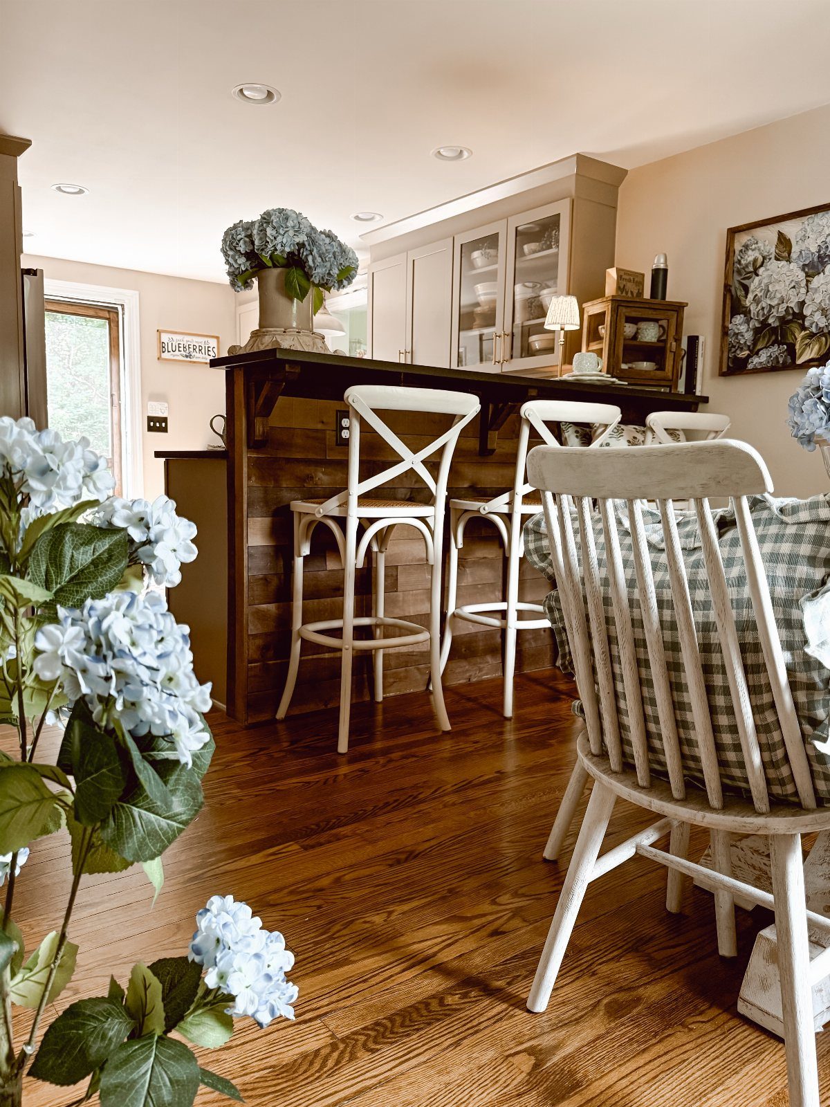 White X-back stools tucked under a dark wood breakfast bar with blue hydrangeas.